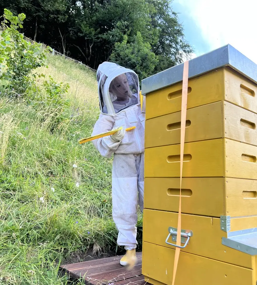 Kind im weissen Imkeranzug mit Stockmeissel neben einem Bienenstock