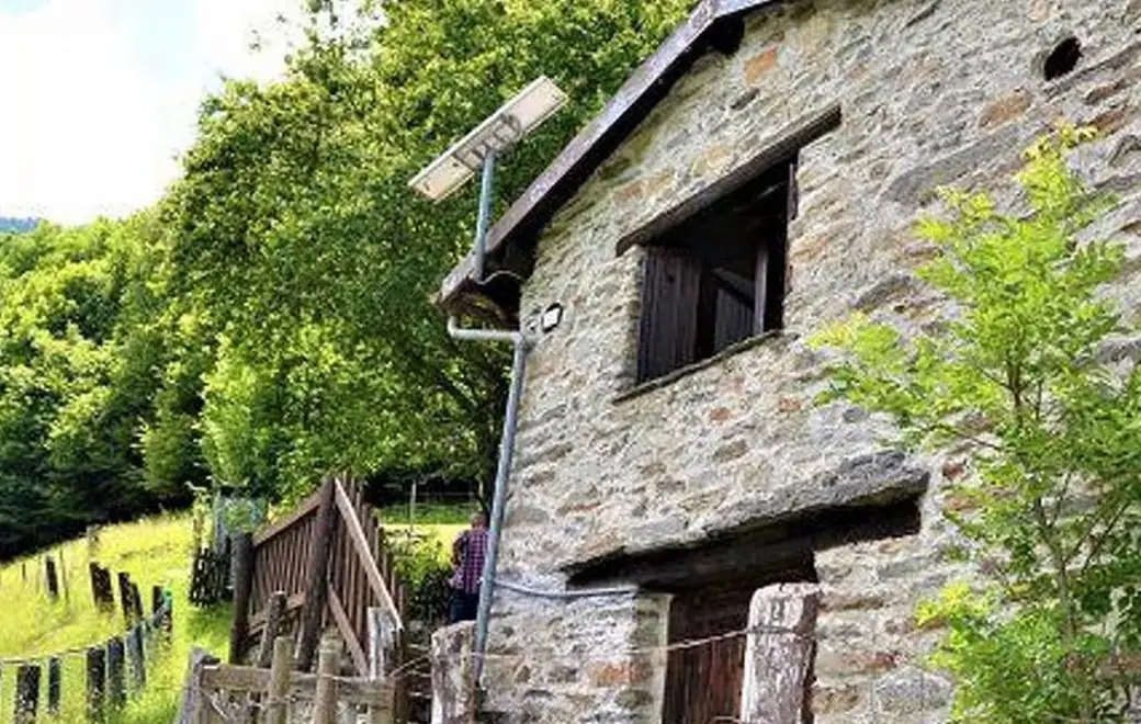 Rustico aus Bruchstein mit Holzfensterläden, davor ein Zaun und die ansteigende Wiese